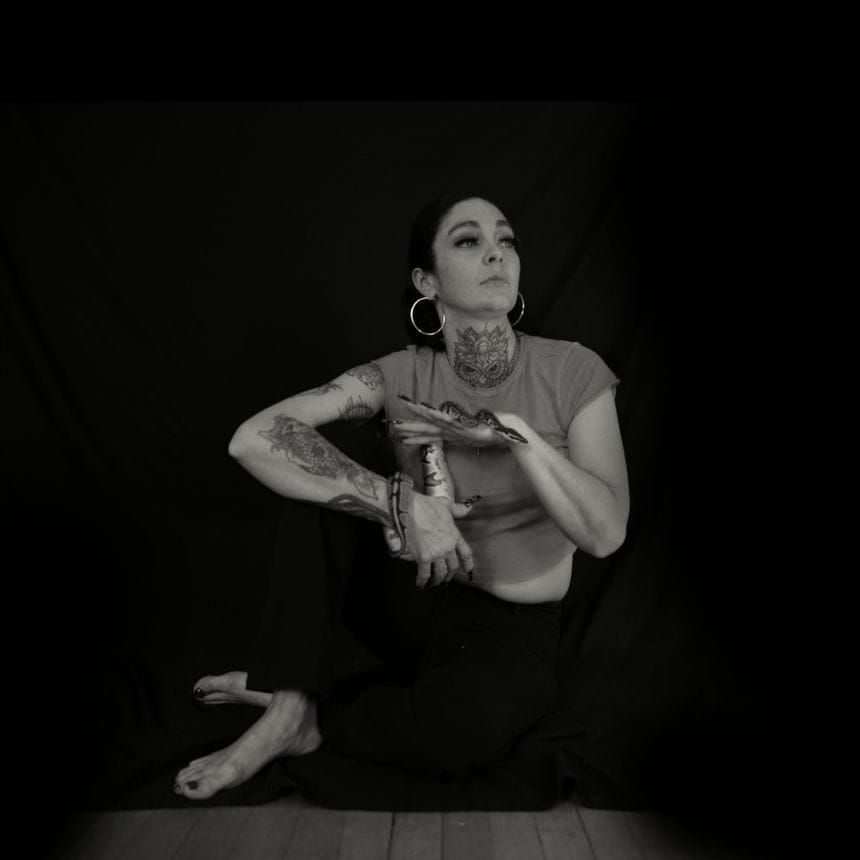 Andrea Martin sits on the floor in front of a dark background with her arm propped up on her right knee. She’s also holding her pet snake, which is wrapped around her right wrist. A younger woman, she wears a red t-shirt and large hoop earrings, and has her dark hair pulled back.