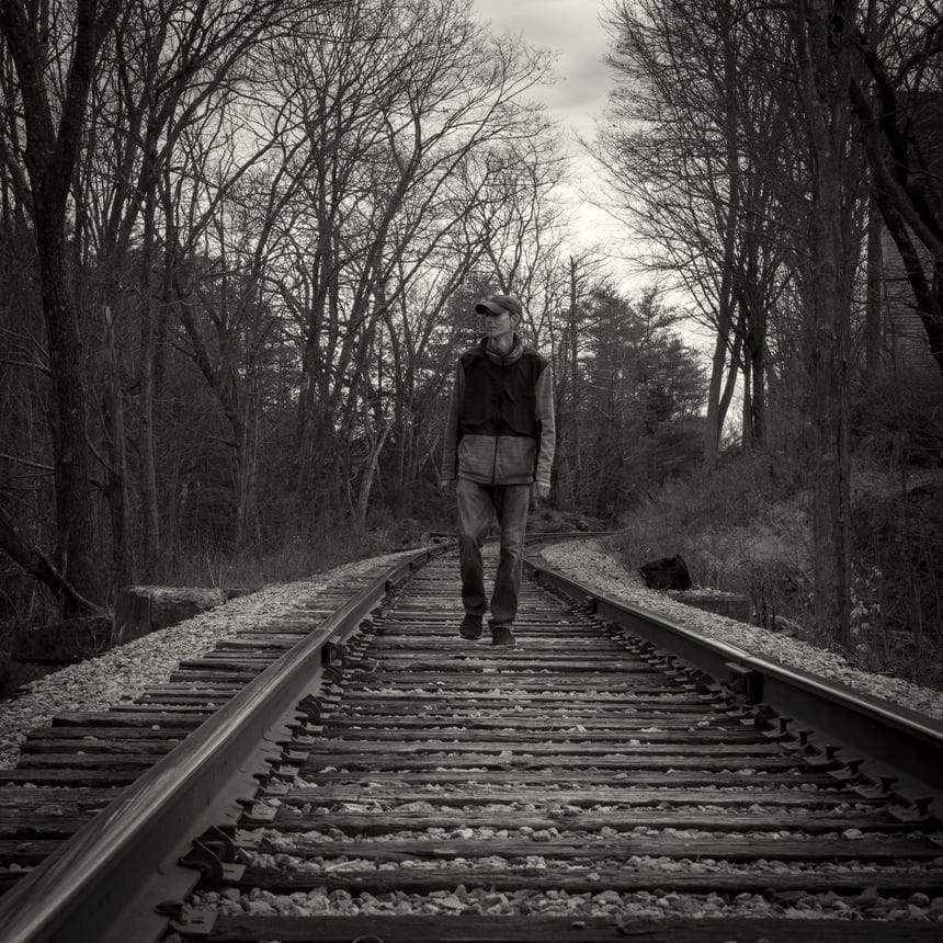 Black-and-white photograph of Andy Perkins, age 48, walking along the railroad tracks in Milton, NH, wearing a baseball hat, zip-up jacket, jeans, and dark sneakers.